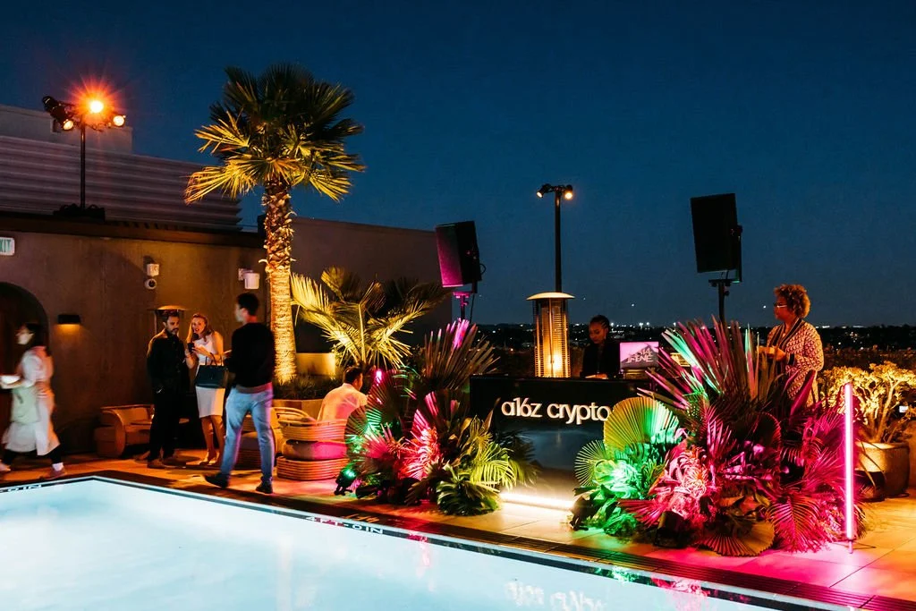 Nighttime rooftop party with people socializing near a pool, illuminated tropical plants, DJ booth with 'albz crypto' sign, palm trees, colorful lighting, city skyline in the background.