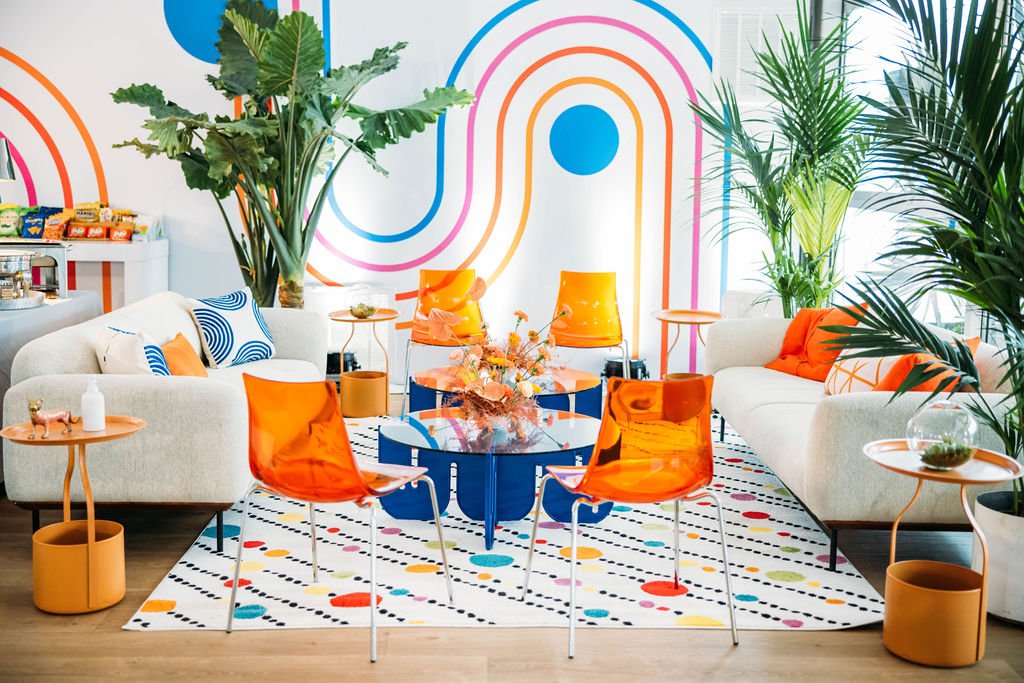 Colorful modern lounge with white sofas, orange chairs, and a striped rug, decorated with large potted plants and abstract wall art.