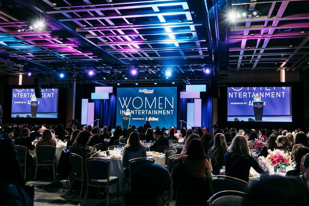 A large conference or awards event with many women seated at round tables, a stage with large screens displaying 'Women in Entertainment', and vibrant lighting in purple and blue tones.