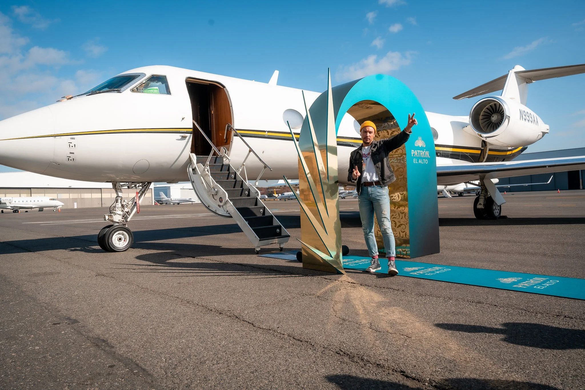 A man standing in front of a private jet airplane at an airport, giving a peace sign, with a decorative opening as part of a promotional display nearby.