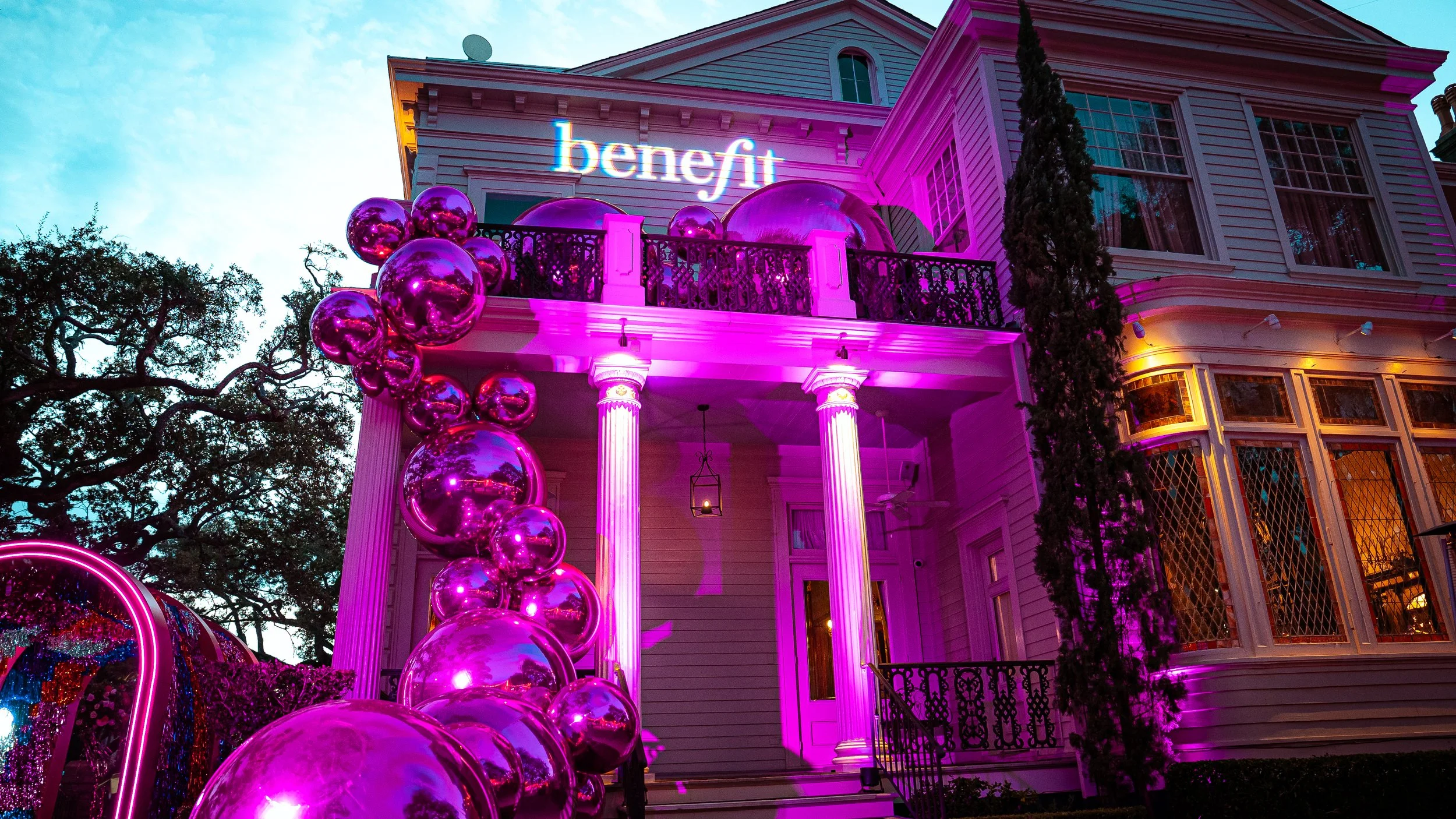 A large white house decorated with purple and pink lighting, featuring a balcony, columns, and a bay window. Purple balloons adorn the front, and there are trees in the background.